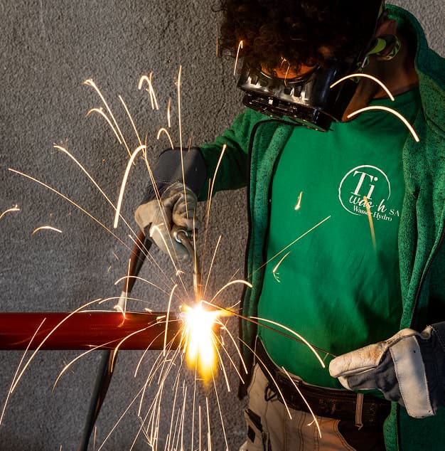 Operatore al lavoro con taglio a fiamma: scintille dorate, grembiule verde e protezioni, sfondo di metallo scuro.