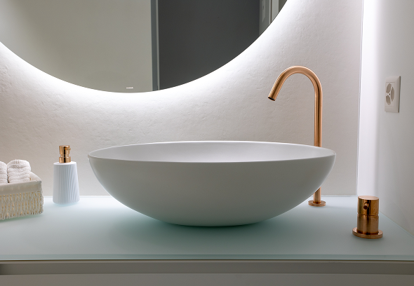 Lavabo ovale bianco su piano vetro chiaro, rubinetto dorato a collo alto, specchio circolare retroilluminato, accessori bagno in ceramica e tessuto coordinato.