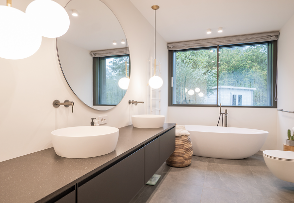 Bagno moderno con doppio lavabo rotondo su piano scuro, grande specchio tondo e vasca freestanding luminosa, vetri grandi e atmosfera luminosa.