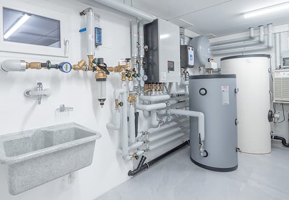 Impianto tecnico con tubature, valvole e serbatoio cilindrico in una sala bianca, sistema di riscaldamento/raffreddamento industriale.