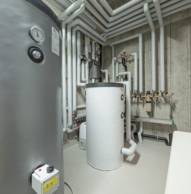 Sala tecnica con serbatoio d’acqua e tubazioni metalliche, valvole e strumenti di controllo su pareti concrete.