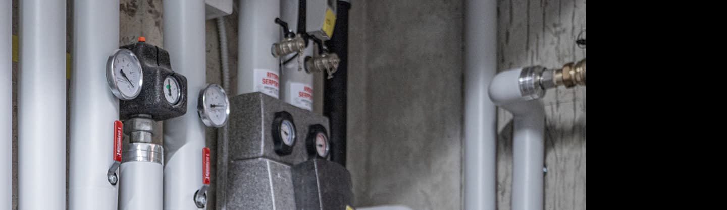Immagine di componenti meccanici e tubature in un ambiente tecnico, con valvole, manometri e tubazioni visibili su sfondo cemento.