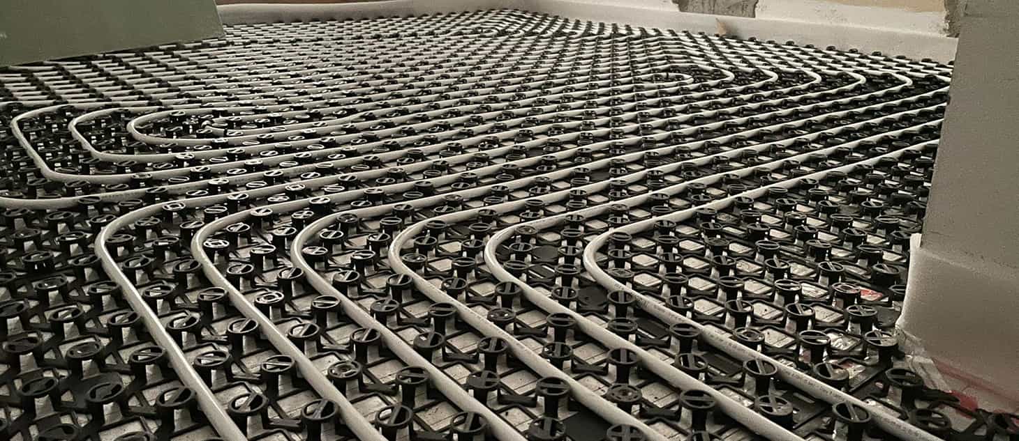 Rete di riscaldamento a pavimento in lastre metalliche con serpentine, vista ravvicinata su superficie grigliata.